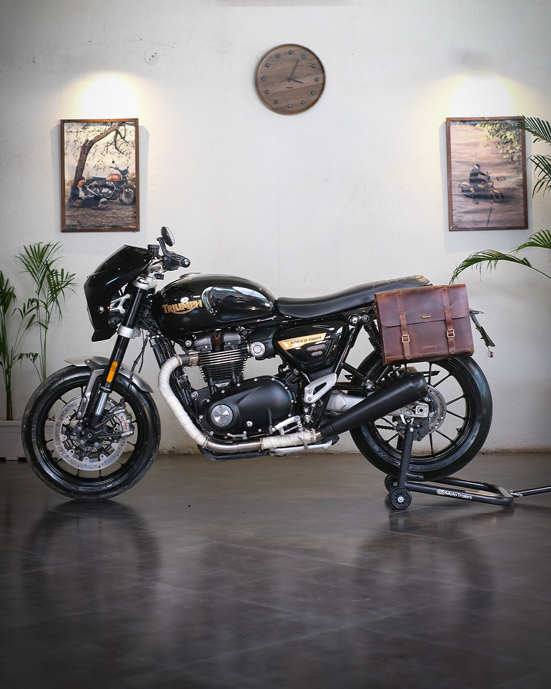 Black motorcycle with a brown leather bag in a room with plants and framed pictures on the wall.