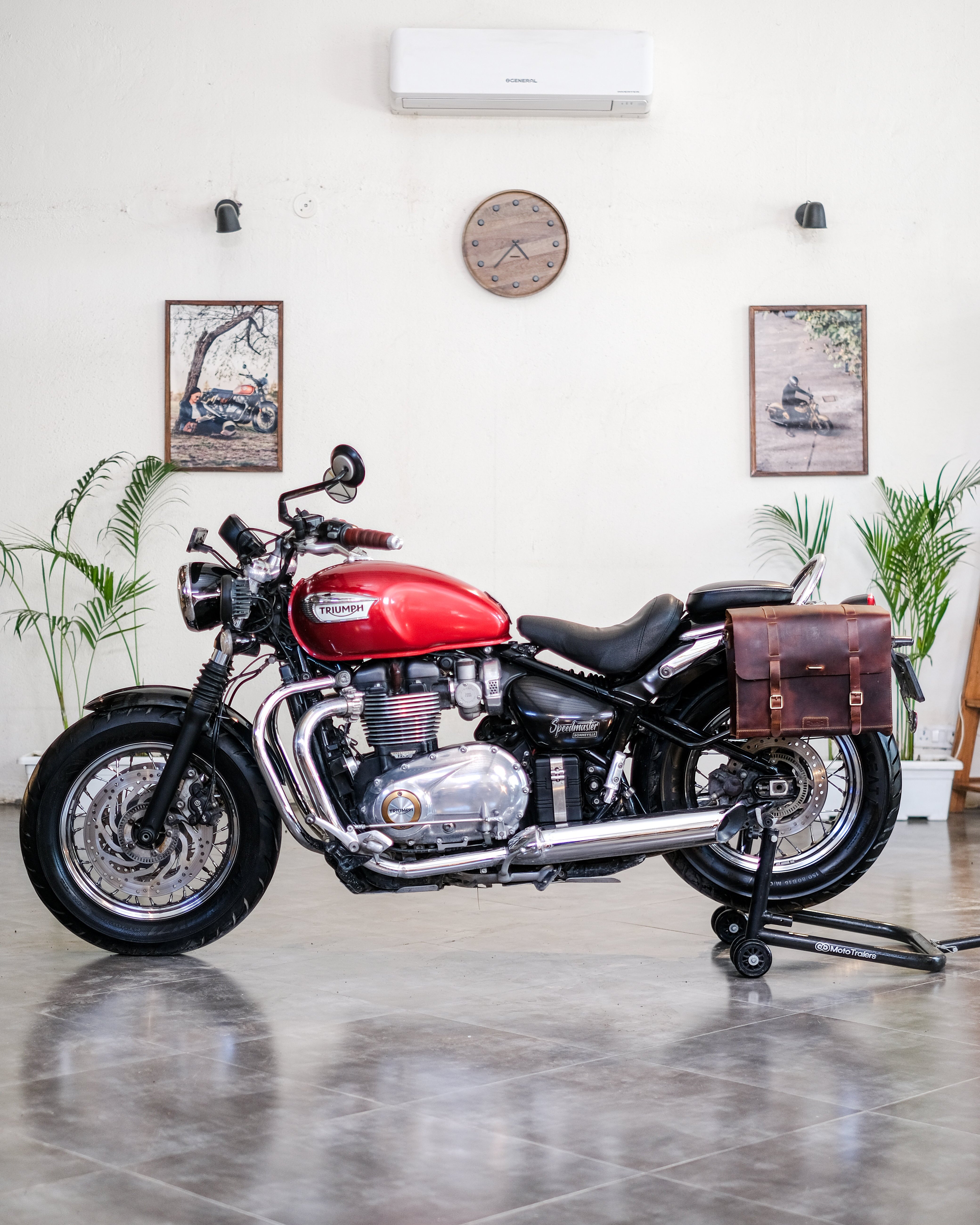 Red motorcycle with a brown leather saddlebag in a room with plants and a clock on the wall.
