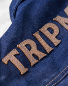 Close-up of a blue denim jacket with 'TRIP' embroidered in brown letters.
